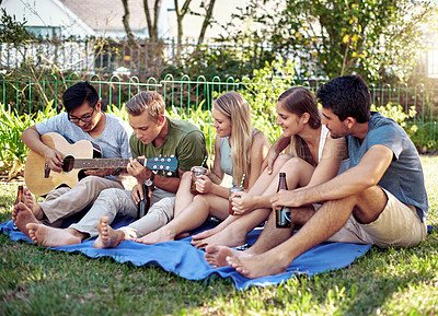 Buy stock photo Full length shot of a young man playing guitar while enjoying a few drinks with his friends outside in the summer sun