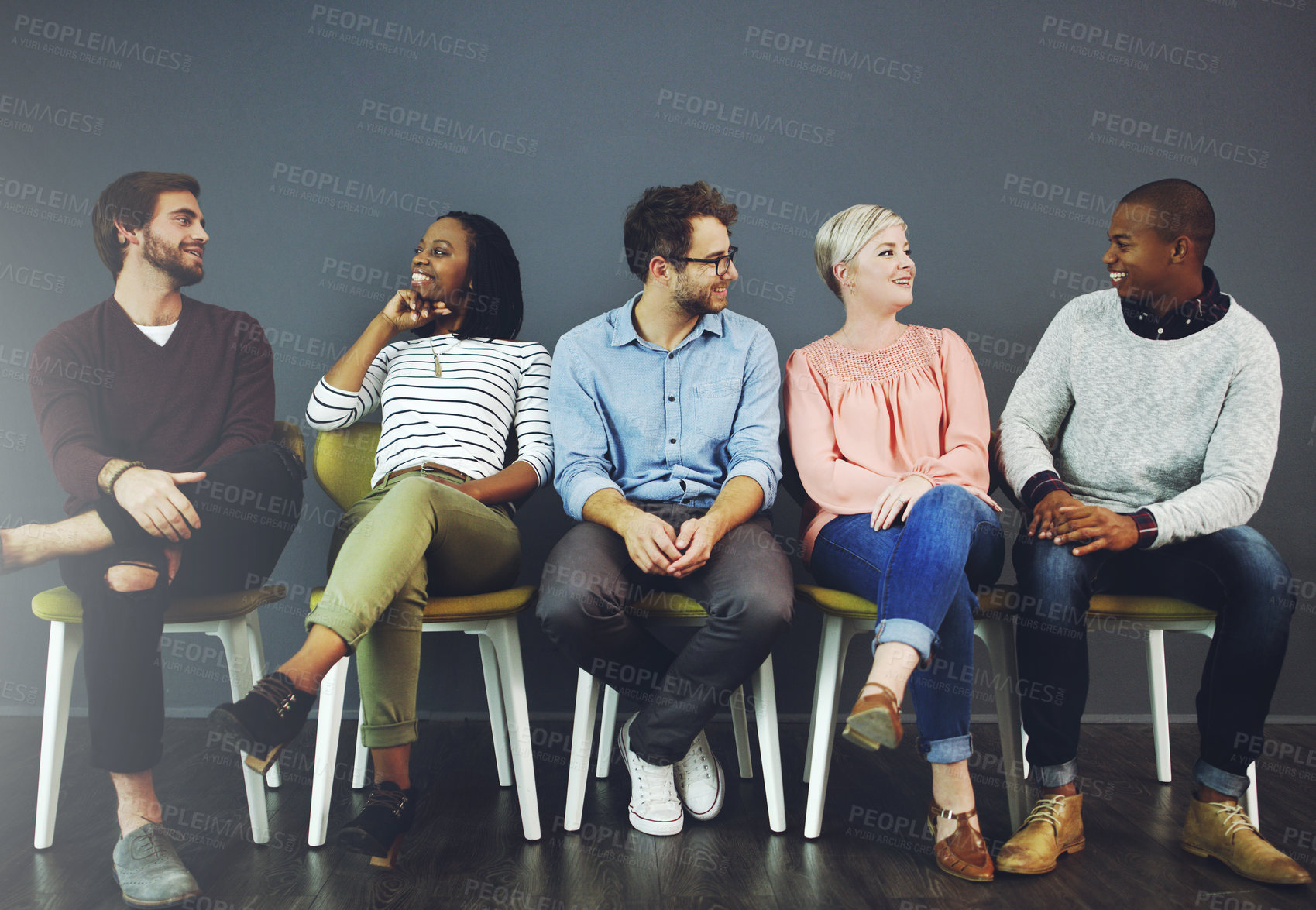 Buy stock photo Studio shot of a group of young people waiting in line on chairs and chatting against a gray background