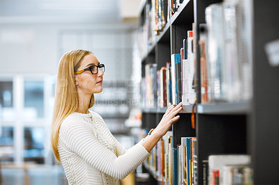 Buy stock photo Shot of a focused young woman taking a book to read from a bookshelf inside of a library