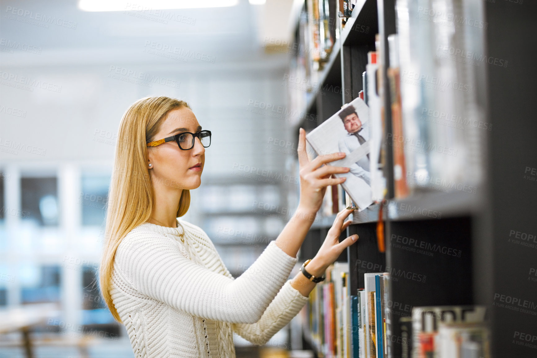 Buy stock photo Shot of a focused young woman taking a book to read from a bookshelf inside of a library