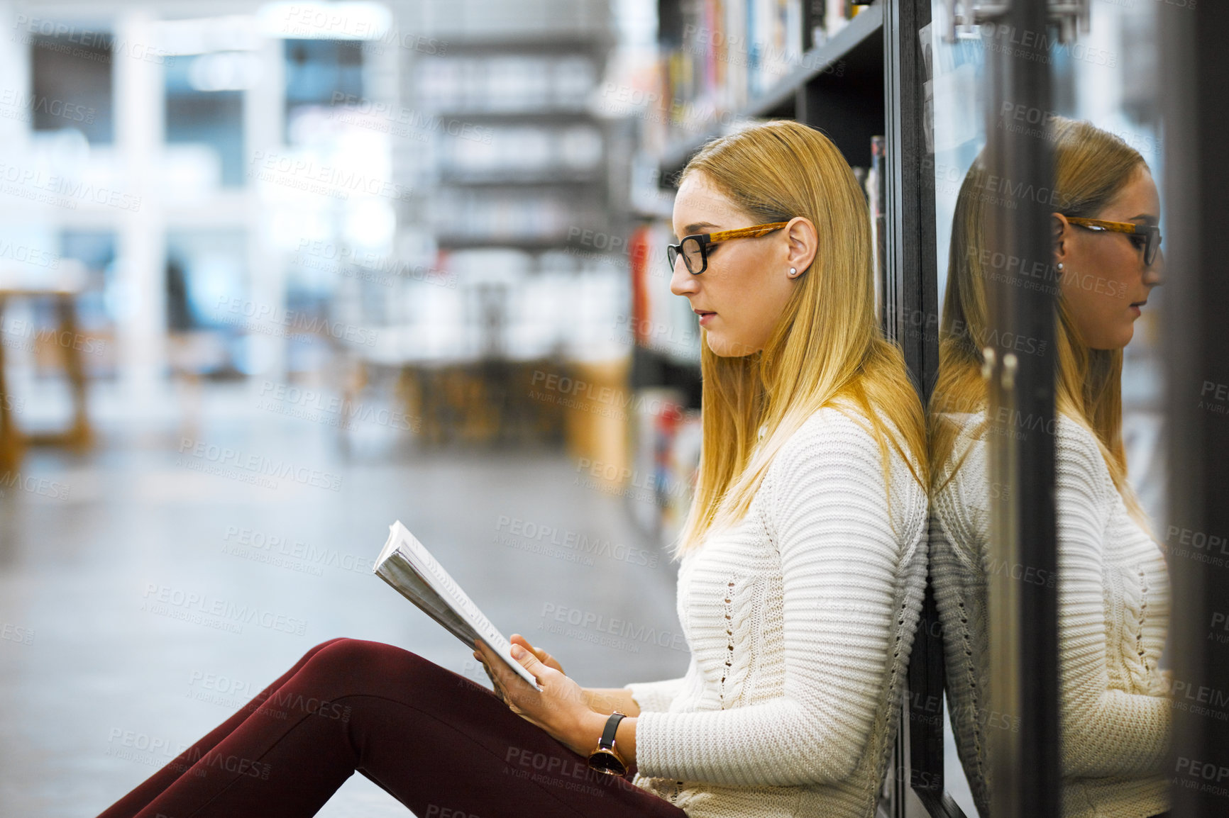 Buy stock photo Shot of a focused young woman reading a book while being seated on the floor in a library