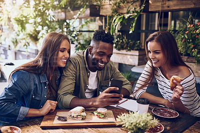Buy stock photo Shot of three friends looking at something on a cellphone while out for lunch