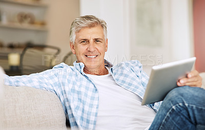 Buy stock photo Shot of a mature man using his digital tablet while relaxing on his sofa at home