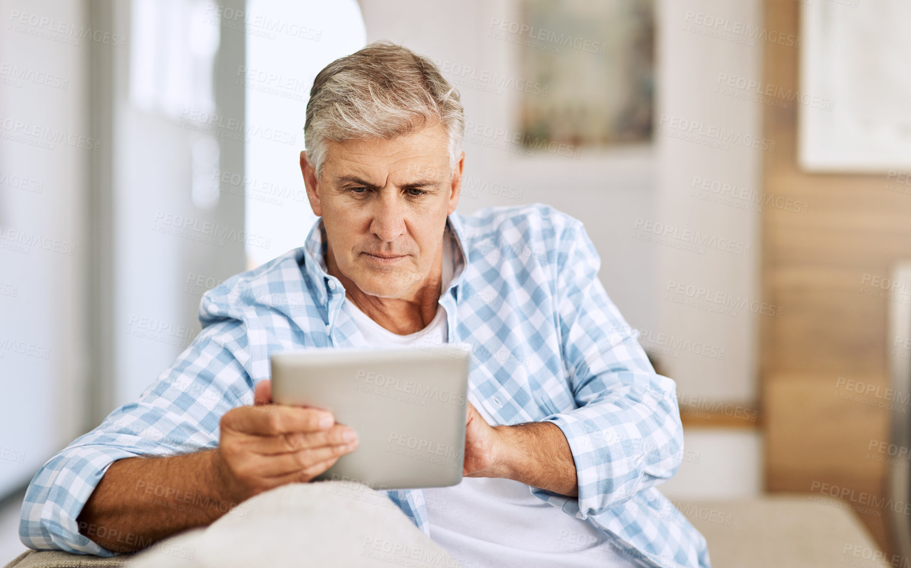 Buy stock photo Shot of a mature man using his digital tablet while relaxing on his sofa at home