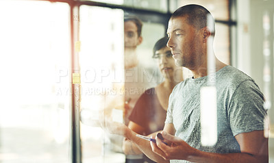 Buy stock photo Cropped shot of a group of young Shot of a man writing ideas on sticky notes with his team behind him planning on a glass board
