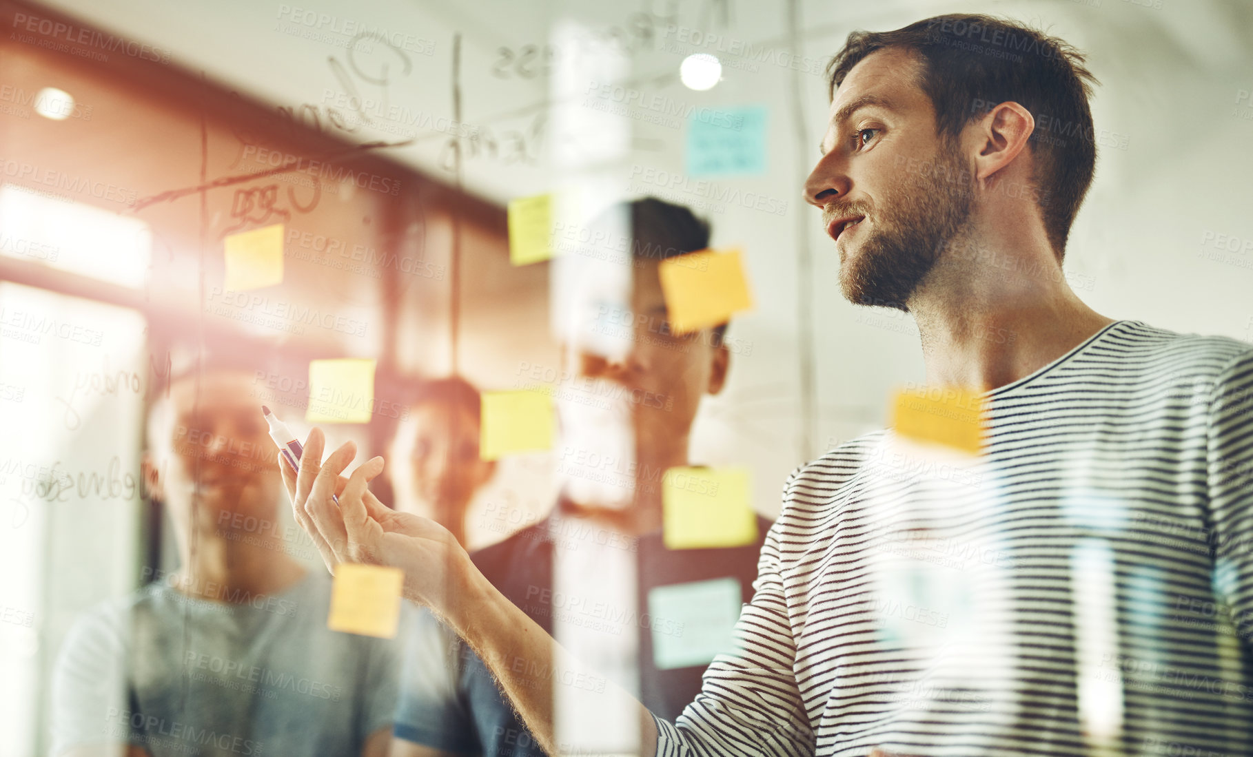 Buy stock photo Group of young designers brainstorm as a team. Writing down new ideas on glass board inside workplace together. Business people meeting at the office to discuss notes and share creative designs.