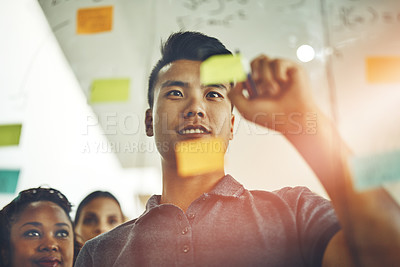 Buy stock photo A marketing professional brainstorming ideas with colleagues, writing on transparent board with sticky notes during meeting. Young, Asian entrepreneur discussing a work project schedule or timeline.