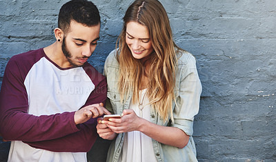 Buy stock photo Shot of a young man and woman standing outdoors and using a mobile phone against a gray wall