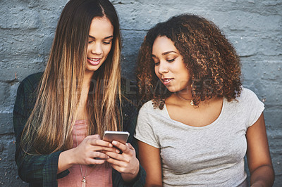 Buy stock photo Shot of two young women standing outdoors and using a mobile phone against a gray wall