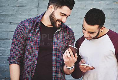 Buy stock photo Shot of two young men standing outdoors and using a mobile phone against a gray wall