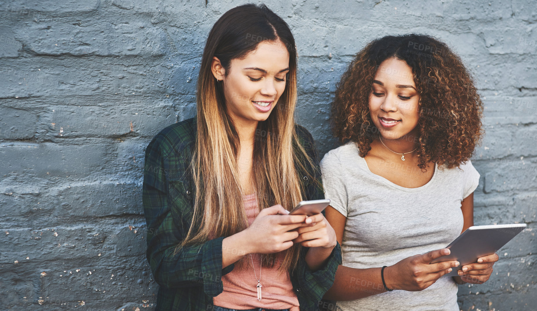 Buy stock photo Shot of two young women standing outdoors and using their digital tablets against a gray wall