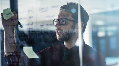 Buy stock photo Shot of a young businessman brainstorming with sticky notes on a glass wall in a modern office