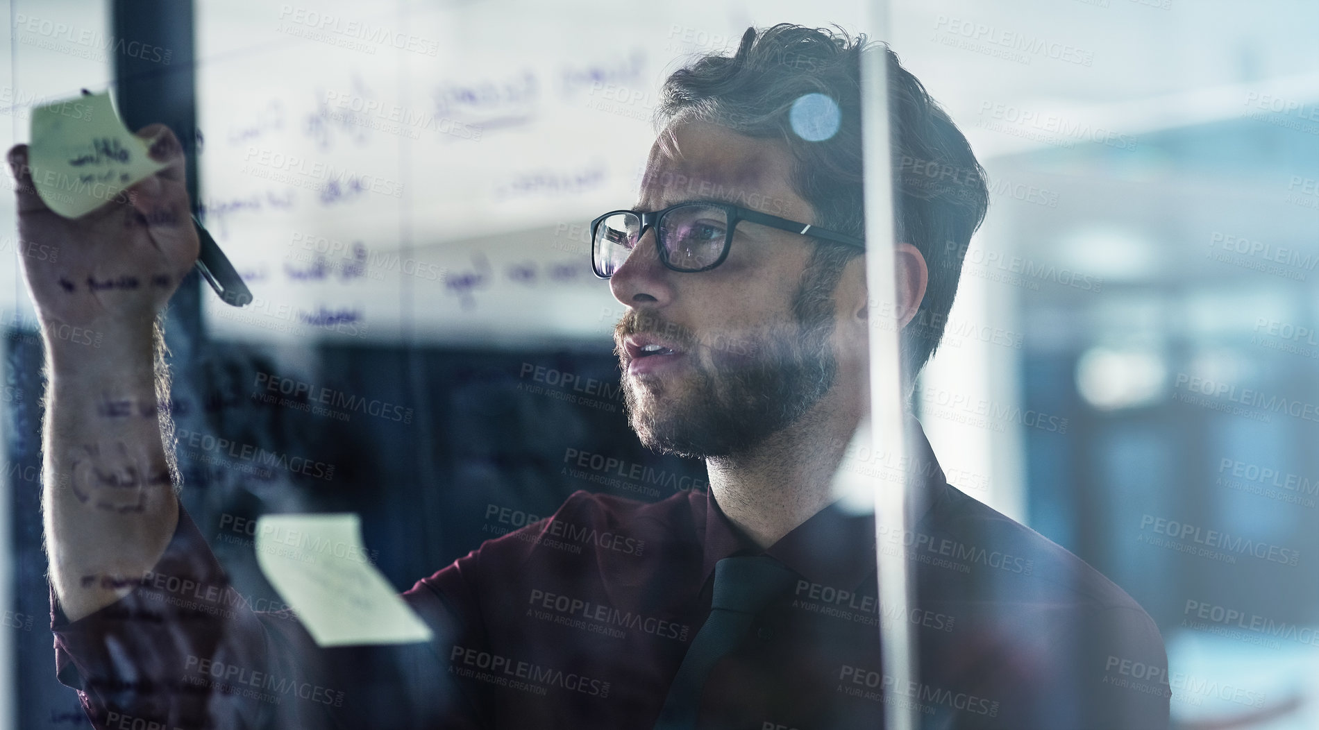 Buy stock photo Shot of a young businessman brainstorming with sticky notes on a glass wall in a modern office