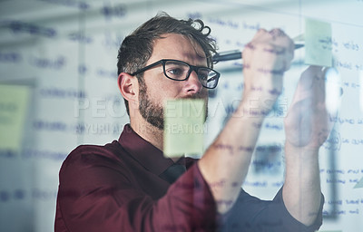 Buy stock photo Shot of a young businessman brainstorming with sticky notes on a glass wall in a modern office