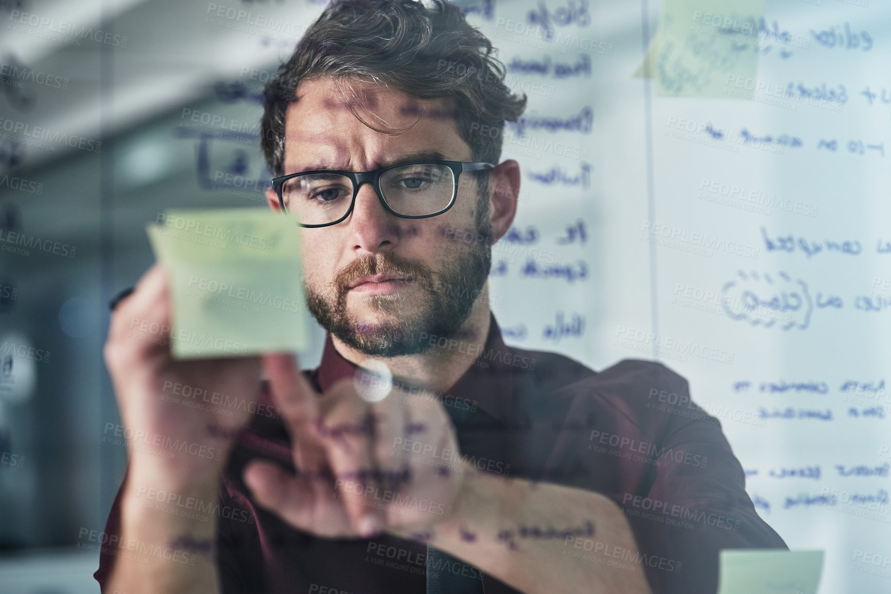 Buy stock photo Shot of a young businessman brainstorming with sticky notes on a glass wall in a modern office