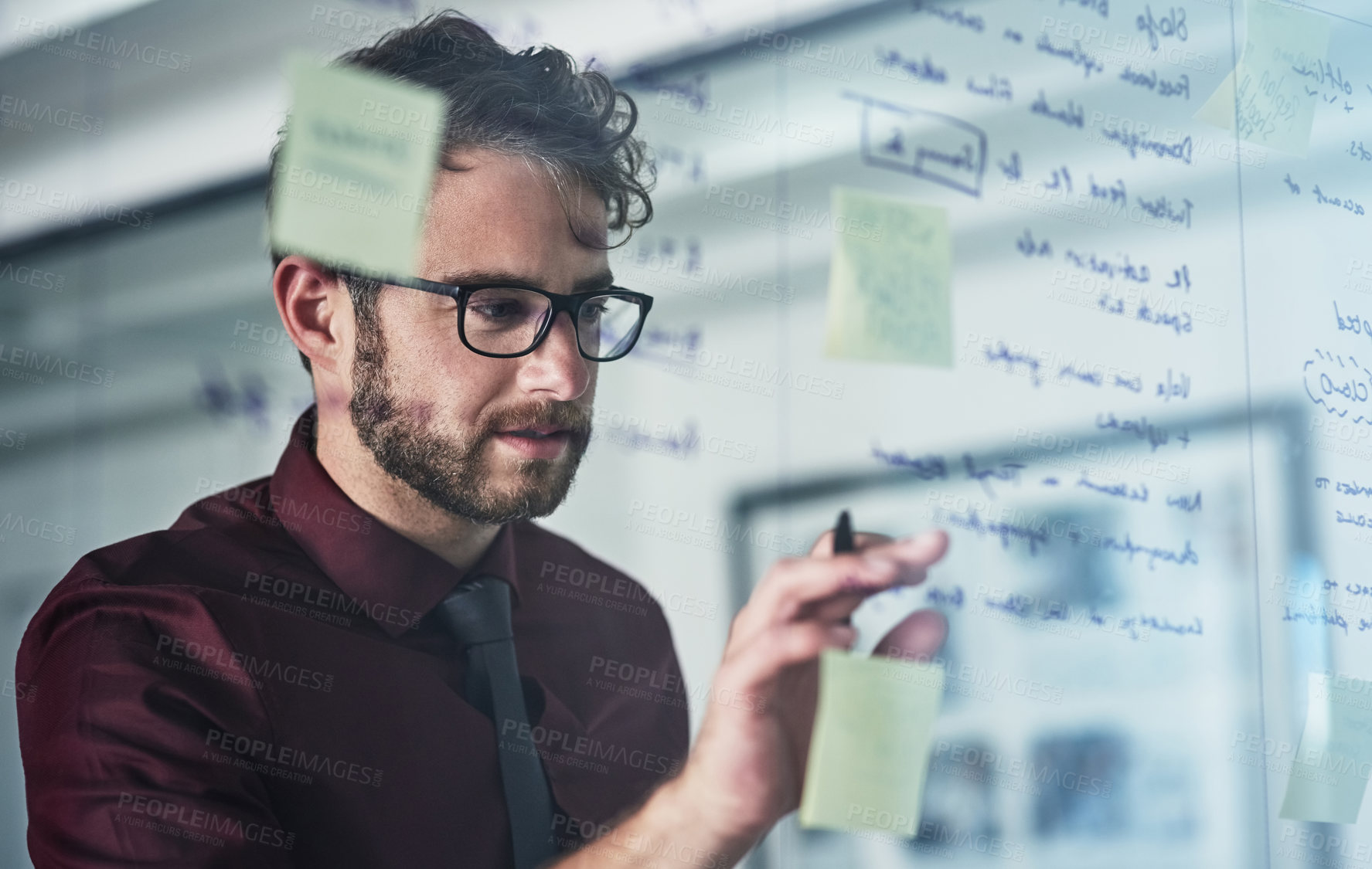 Buy stock photo Shot of a young businessman brainstorming with sticky notes on a glass wall in a modern office