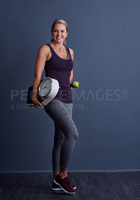 Buy stock photo Studio portrait of an attractive mature woman holding an apple and a weightscale against a blue background
