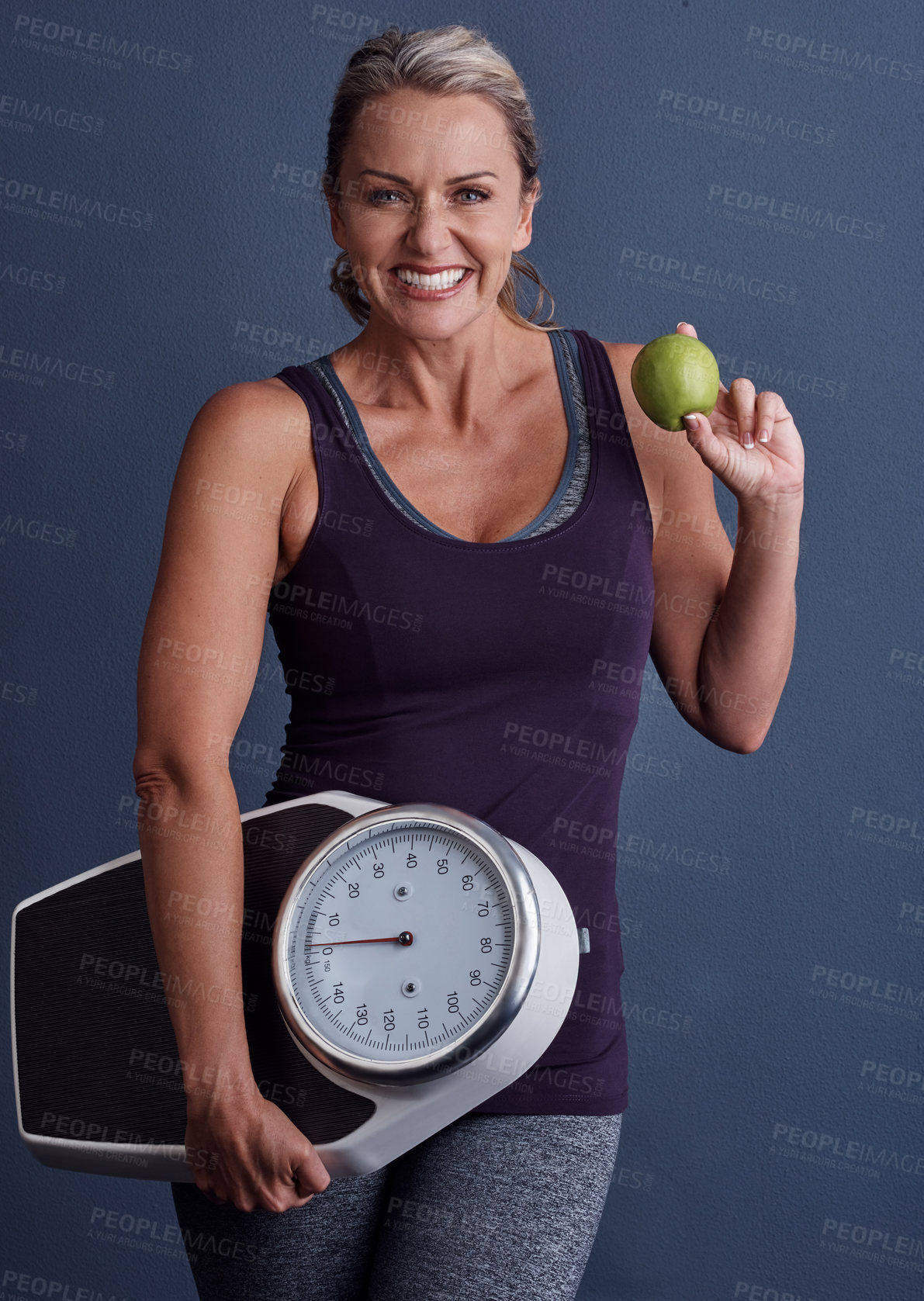 Buy stock photo Studio portrait of an attractive mature woman holding an apple and a weightscale against a blue background