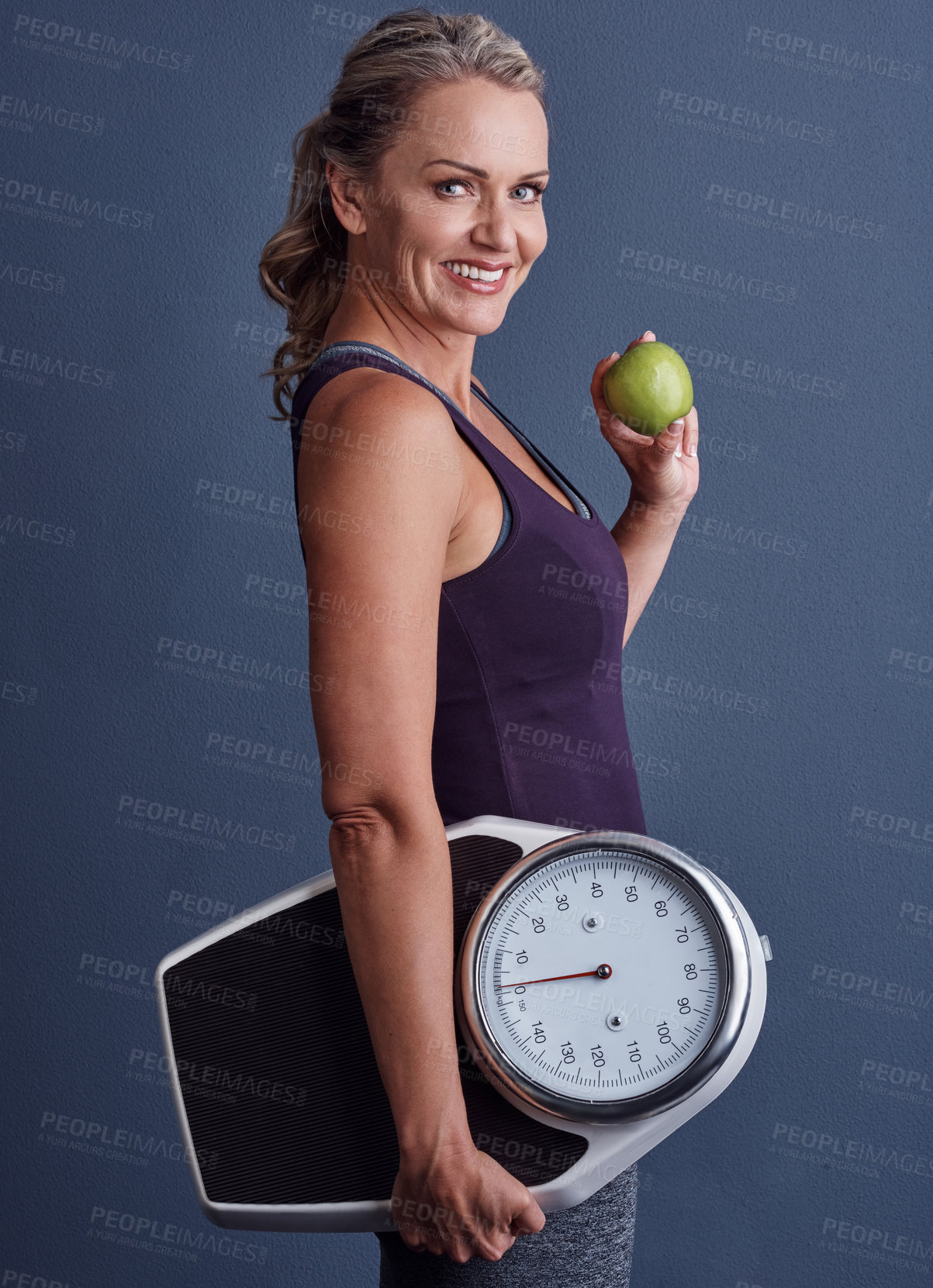 Buy stock photo Studio portrait of an attractive mature woman holding an apple and a weightscale against a blue background