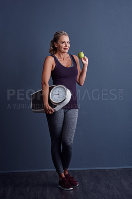 Buy stock photo Studio portrait of an attractive mature woman holding an apple and a weightscale against a blue background