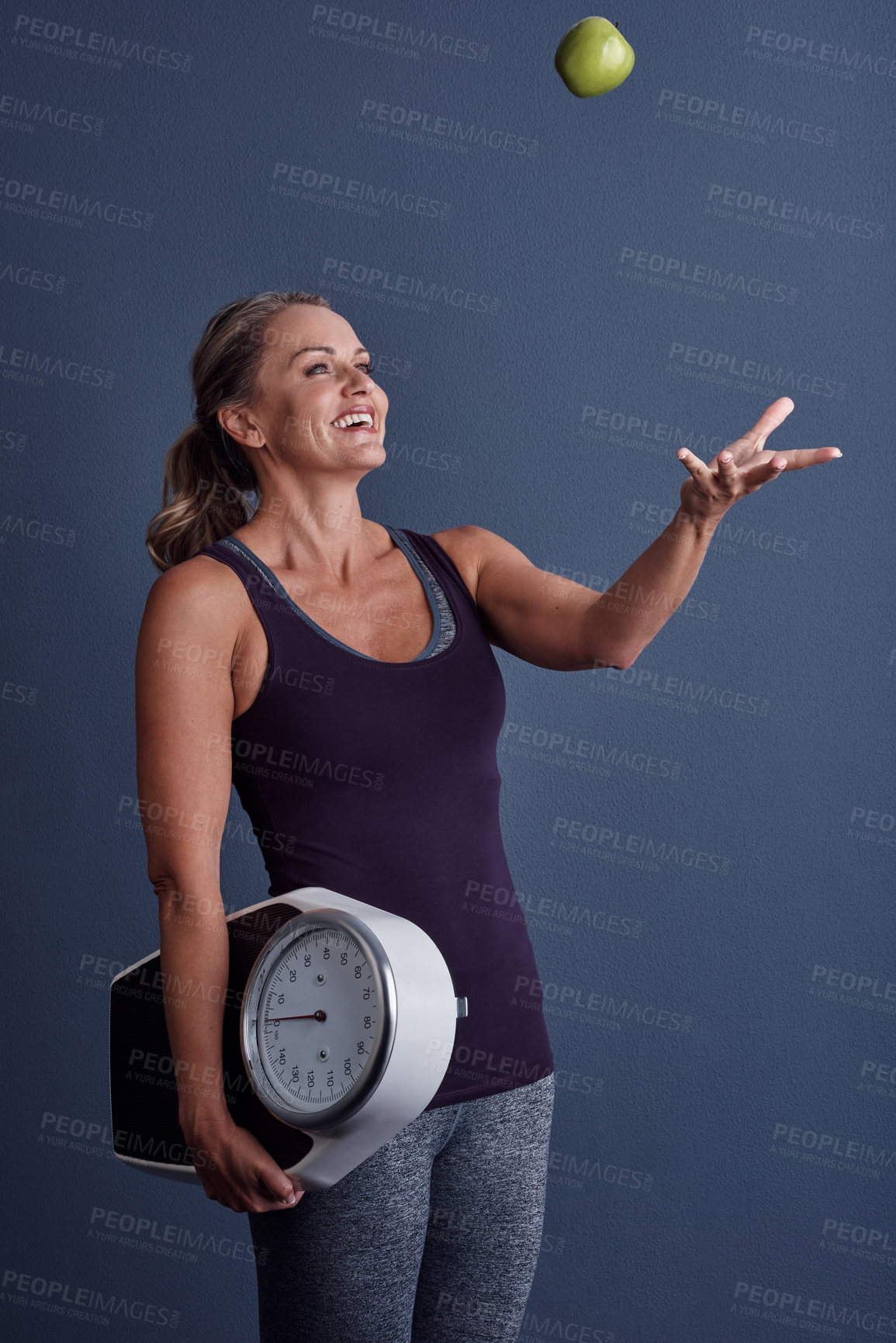 Buy stock photo Studio shot of an attractive mature woman throwing an apple and holding a weightscale against a blue background