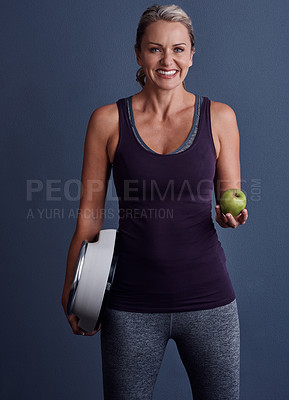 Buy stock photo Studio portrait of an attractive mature woman holding an apple and a weightscale against a blue background