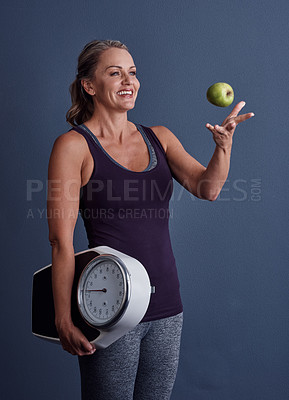 Buy stock photo Studio shot of an attractive mature woman throwing an apple and holding a weightscale against a blue background