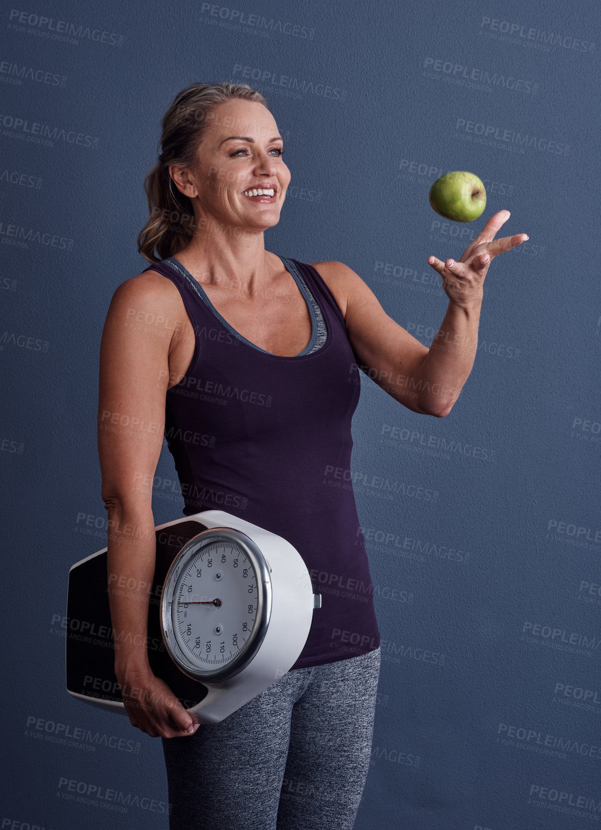 Buy stock photo Studio shot of an attractive mature woman throwing an apple and holding a weightscale against a blue background