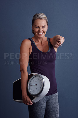 Buy stock photo Studio portrait of an attractive mature woman holding a weightscale and pointing at the camera against a blue background