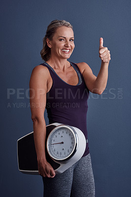 Buy stock photo Studio portrait of an attractive mature woman holding a weightscale and giving thumbs up against a blue background