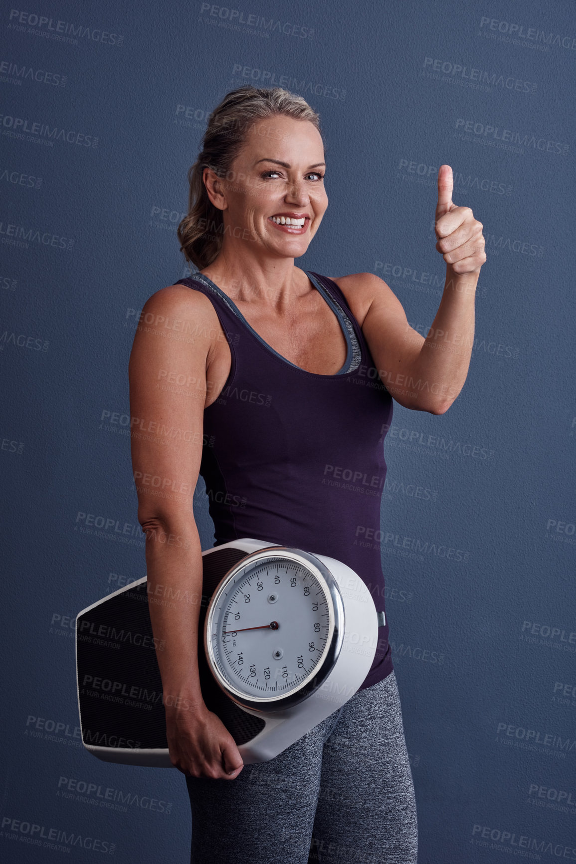 Buy stock photo Studio portrait of an attractive mature woman holding a weightscale and giving thumbs up against a blue background