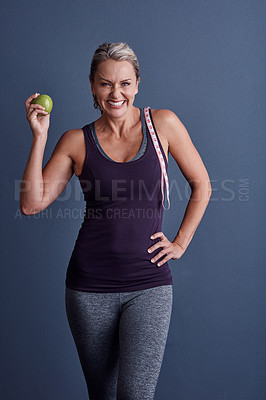 Buy stock photo Studio portrait of an attractive mature woman holding an apple with a tape measure over her shoulder against a blue background
