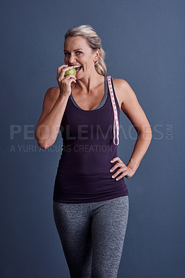 Buy stock photo Studio portrait of an attractive mature woman biting into an apple with a tape measure over her shoulder against a blue background