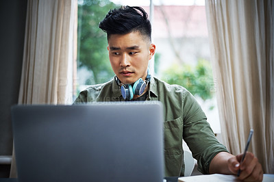 Buy stock photo Cropped shot of a handsome young man taking notes while working on his laptop at home