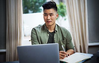 Buy stock photo Cropped shot of a handsome young man taking notes while working on his laptop at home