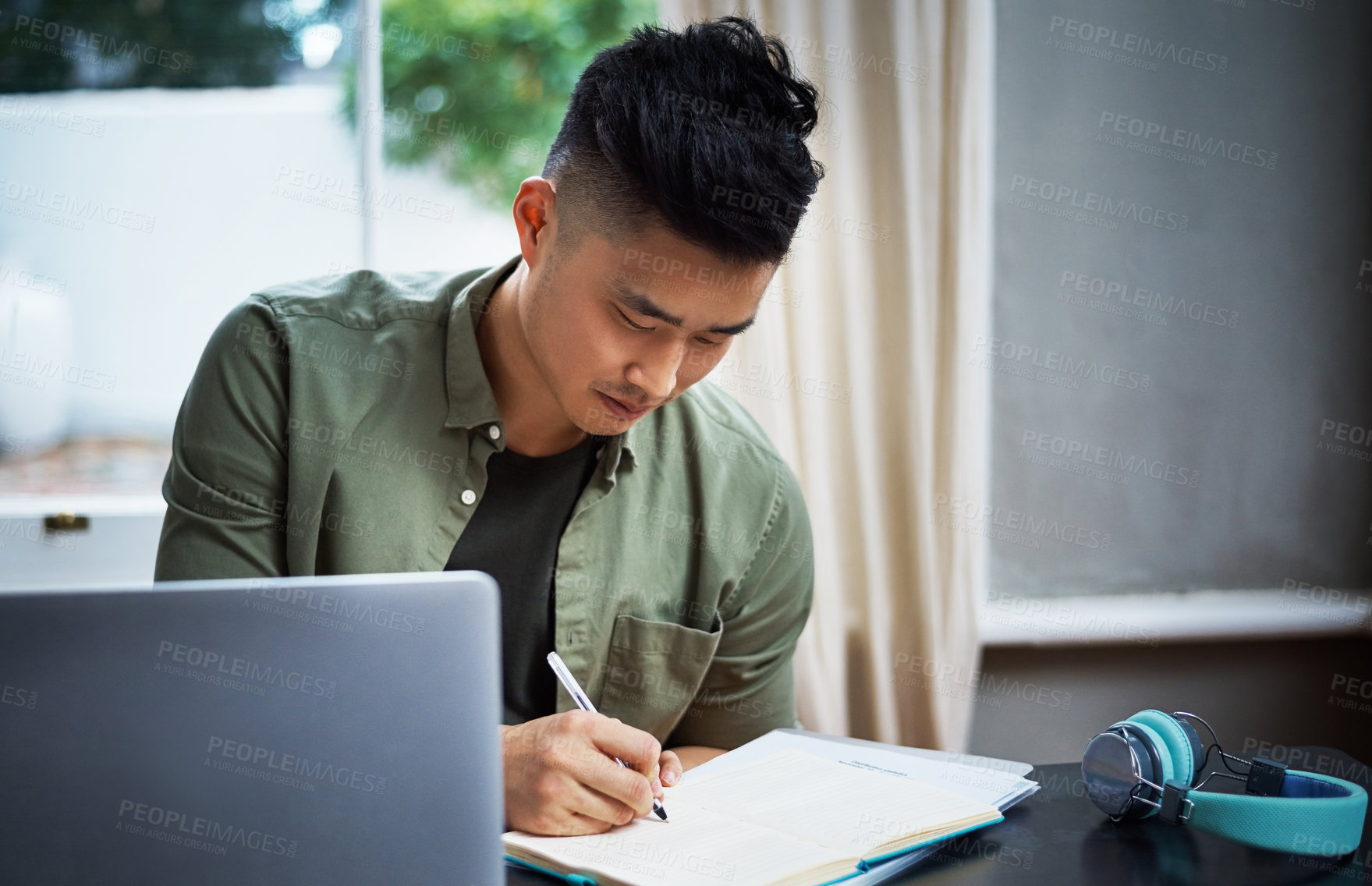 Buy stock photo Cropped shot of a handsome young man taking notes while working on his laptop at home