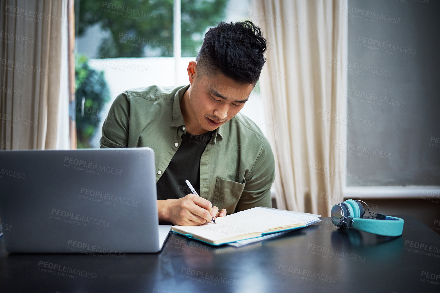 Buy stock photo Cropped shot of a handsome young man taking notes while working on his laptop at home