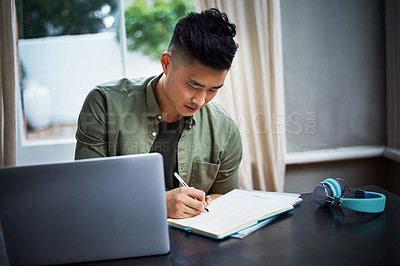 Buy stock photo Cropped shot of a handsome young man taking notes while working on his laptop at home