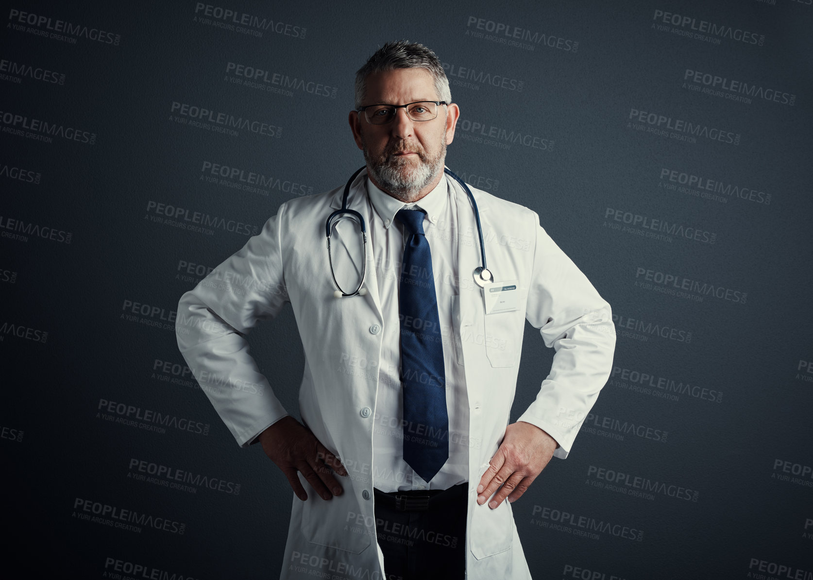 Buy stock photo Studio portrait of a handsome mature male doctor standing with his hands on his hips against a dark background