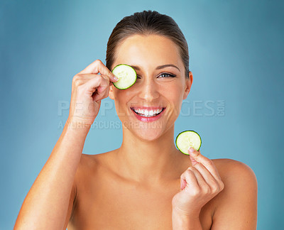 Buy stock photo Studio portrait of a beautiful young woman covering her eye with a cucumber slice against a blue background