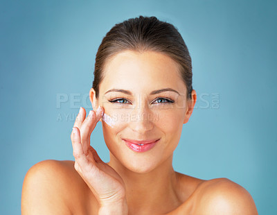 Buy stock photo Studio portrait of a beautiful young woman applying moisturizer to her face against a blue background