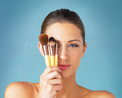 Buy stock photo Studio portrait of a beautiful young woman holding makeup brushes in front of her eyes against a blue background