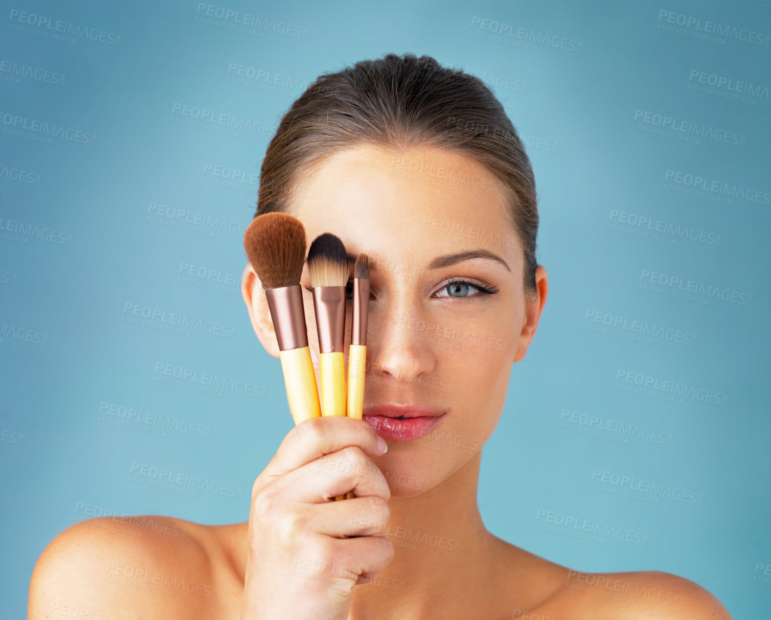 Buy stock photo Studio portrait of a beautiful young woman holding makeup brushes in front of her eyes against a blue background