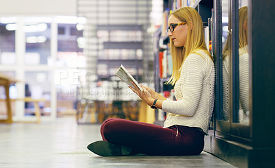 Buy stock photo Full length shot of an attractive young university reading a book while sitting on the library floor