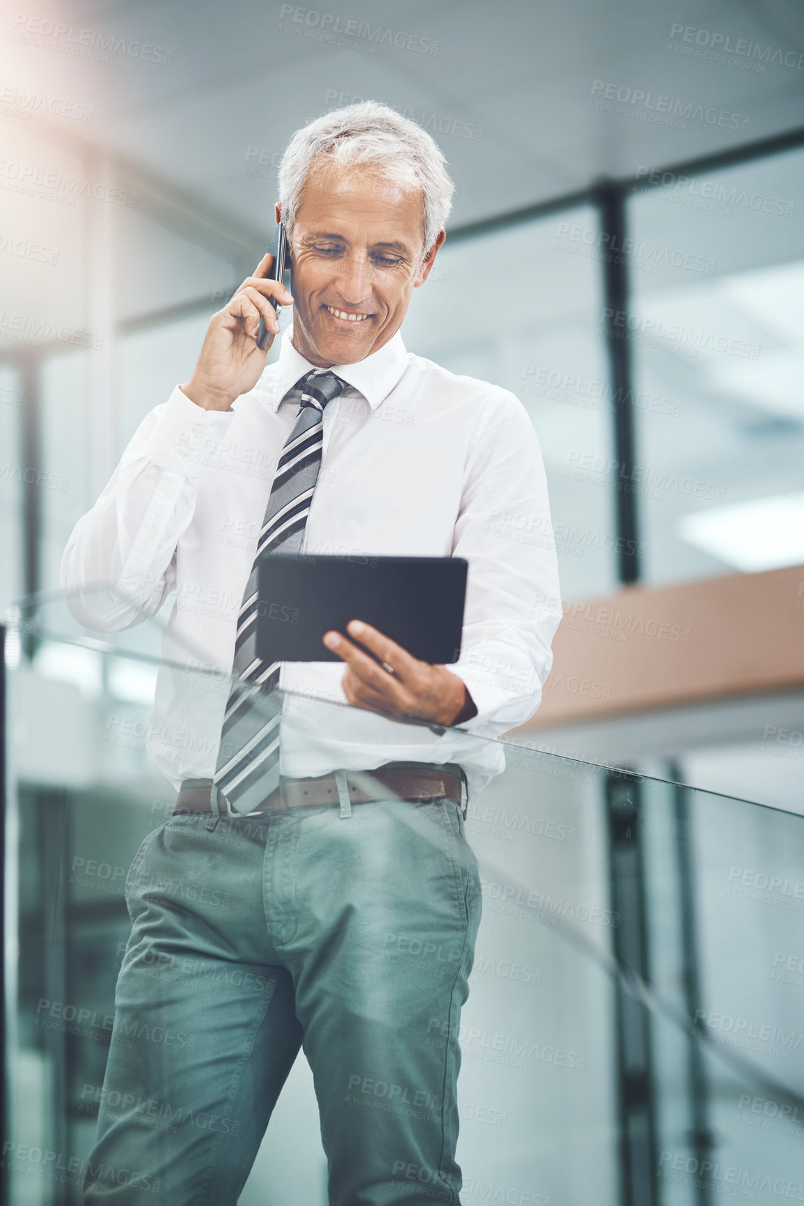 Buy stock photo Cropped shot of a handsome mature businessman making a phonecall and looking at his tablet while standing in the office