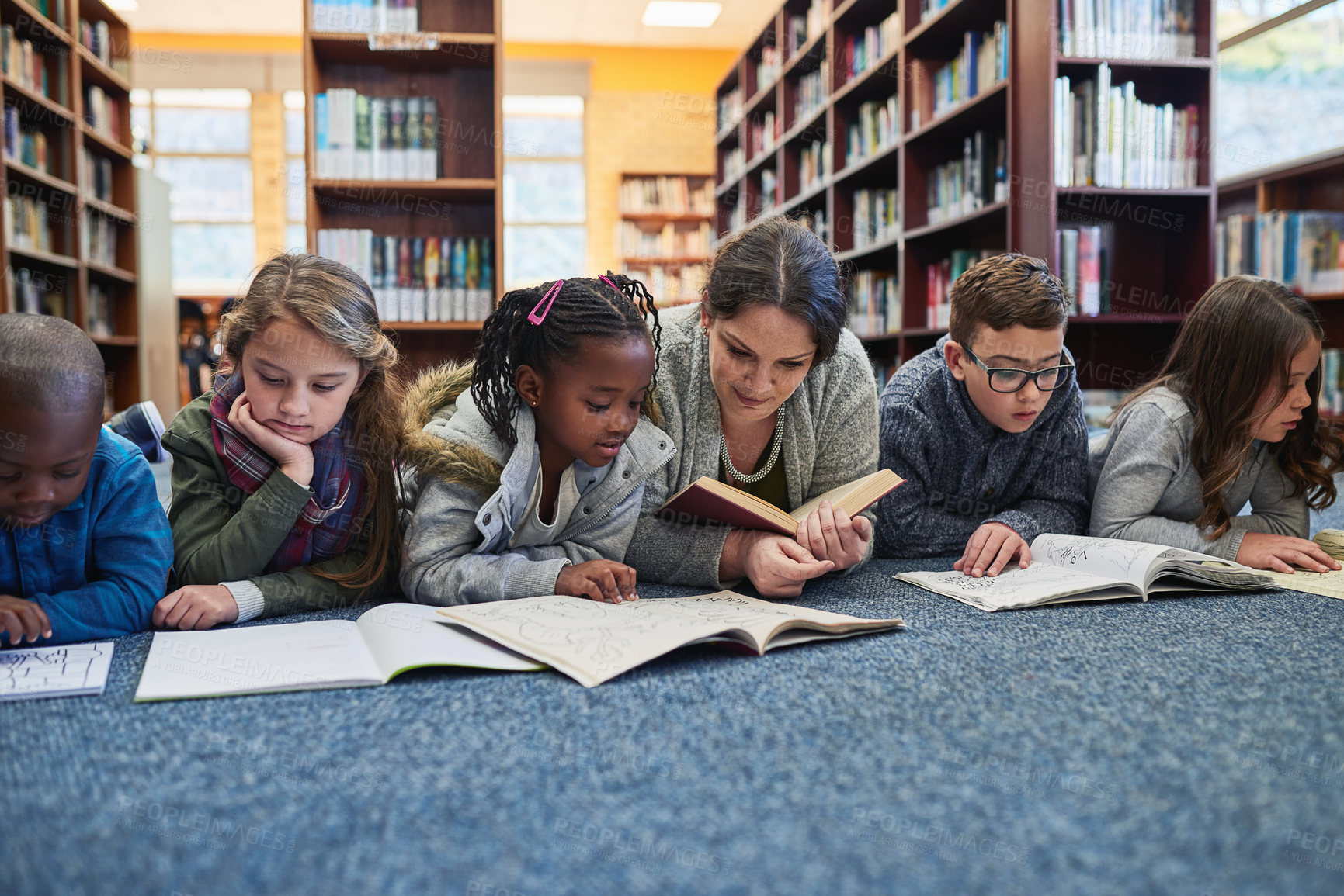 Buy stock photo Education, teacher with children in library and lying on the floor of elementary school. Studying or knowledge with books, diversity and young students with woman reading textbook or notebook.