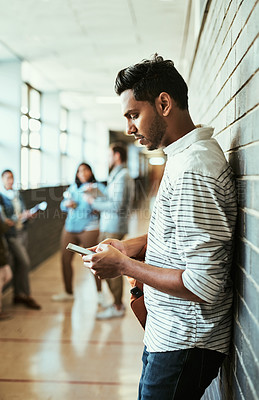 Buy stock photo Cropped shot of a handsome young male university student using his cellphone while standing in a campus corridor