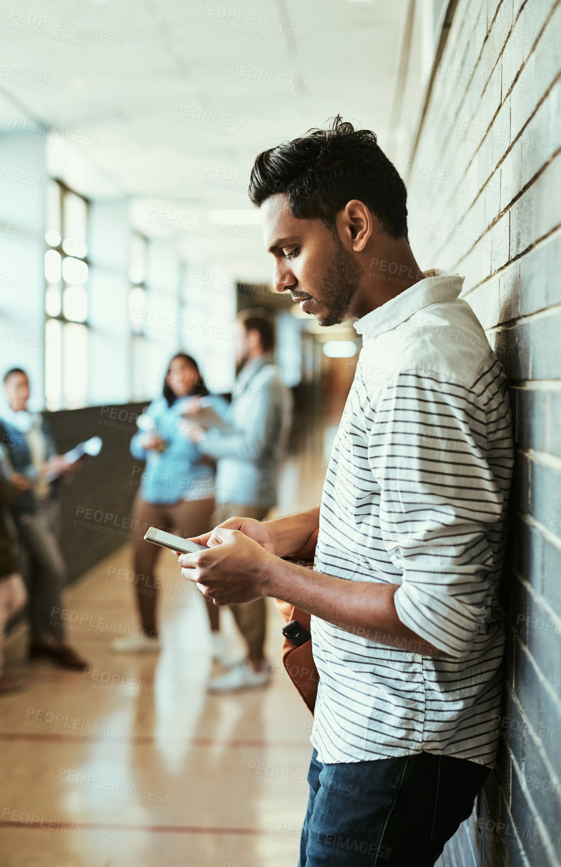 Buy stock photo Cropped shot of a handsome young male university student using his cellphone while standing in a campus corridor