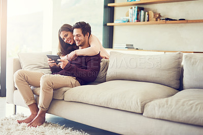 Buy stock photo Shot of a cheerful young couple using a digital tablet while relaxing on the sofa at home