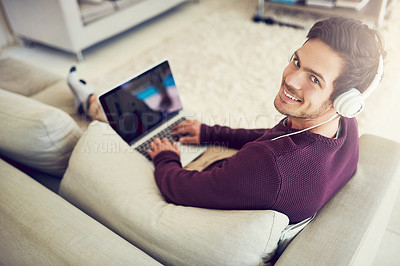 Buy stock photo High angle portrait of a young man listening to music on his laptop while sitting on his sofa and looking into the camera at home